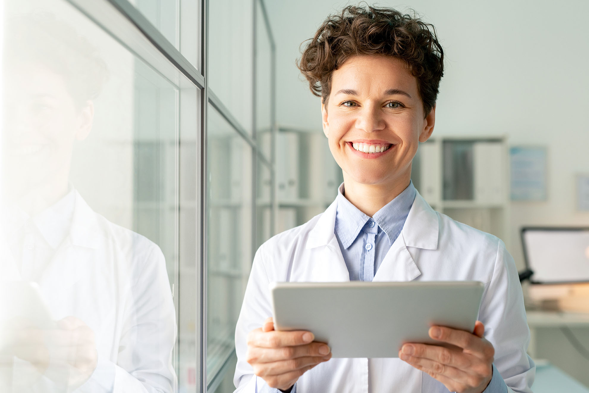 Young pretty successful researcher in whitecoat looking at you with toothy smile while using digital tablet in scientific laboratory by glass wall